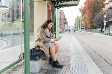 Üppige Frau, die ein Mobiltelefon benutzt, während sie an der Straßenbahnhaltestelle wartet - MEUF04810
