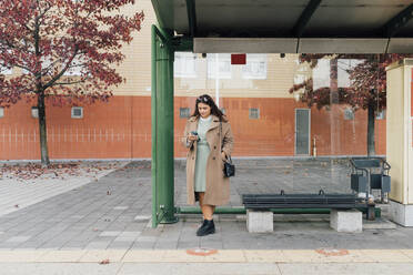 Plus size woman with sling bag using smart phone at bus station - MEUF04826