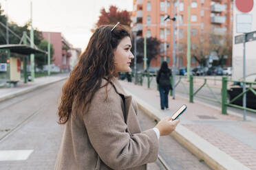 Junge Frau im Mantel schaut weg, während sie ein Mobiltelefon hält - MEUF04829