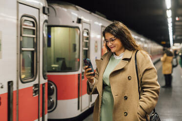 Lächelnde Frau im Mantel, die auf dem Bahnhof ein Mobiltelefon benutzt - MEUF04839