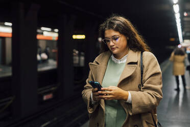 Junge Pendlerin, die am Bahnsteig ein Mobiltelefon benutzt - MEUF04841