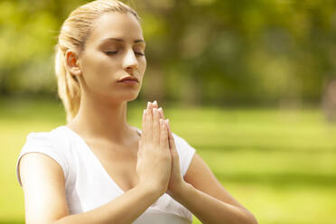 Beautiful blond woman with hands clasped meditating at park - MOMF00960