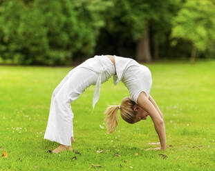 Flexible blond woman performing wheel pose in park - MOMF00964