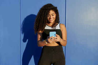 Smiling athlete using mobile phone in front of blue wall - KIJF04302