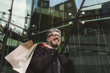 Smiling businessman with shopping bags by glass building - IHF00626