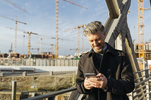 Lächelnder Geschäftsmann mit Mobiltelefon auf einer Brücke - IHF00634