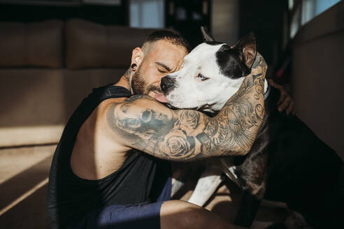 Hipster man with tattooed hand embracing dog at home - MIMFF00725