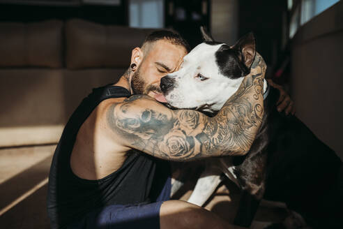Hipster-Mann mit tätowierter Hand umarmt Hund zu Hause - MIMFF00725