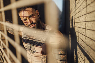 Shirtless man looking through metal grate on sunny day - MIMFF00741