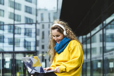 Smiling woman with headphones using laptop in city - IHF00656