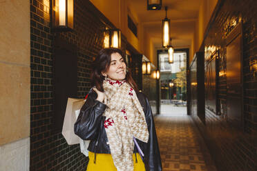 Smiling woman holding shopping bags at corridor - IHF00679