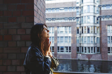 Woman with hands clasped and eyes closed by brick wall - IHF00687