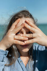 Young female covering face with hands while looking at camera through fingers on on coastline on sunny summer day - ADSF31913