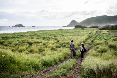 Back view of unrecognizable friends musicians walking with guitars on pathway among green grass on coast near ocean in daytime - ADSF31917