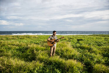 Ruhiger Mann Musiker in legerer Kleidung stehend im grünen Gras an der Küste des Ozeans und spielt akustische Gitarre im Sommer bei Tageslicht - ADSF31923