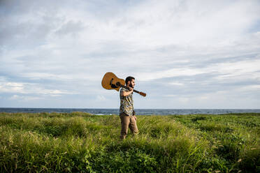 Ruhiger Mann Musiker in Freizeitkleidung stehend mit Akustikgitarre auf der Schulter zwischen grünem Gras an der Küste des Ozeans im Sommer im Tageslicht - ADSF31924