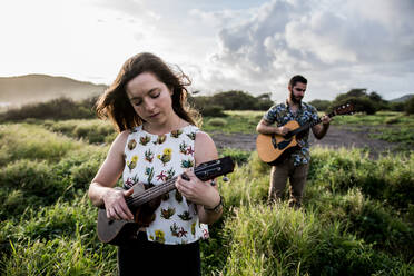 Ruhige Frau in Freizeitkleidung spielt Ukulele und steht im grünen Feld gegen konzentrierte Freund spielt Gitarre in der Natur in sonnigen Tag - ADSF31926