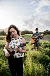 Ruhige Frau in Freizeitkleidung spielt Ukulele und steht im grünen Feld gegen konzentrierte Freund spielt Gitarre in der Natur in sonnigen Tag - ADSF31927