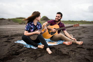 In voller Länge von lächelnden Mann spielt akustische Gitarre mit positiven weiblichen Freund spielt Ukulele beim Sitzen auf sandigen Küste in der Natur in bewölkten Tag - ADSF31931
