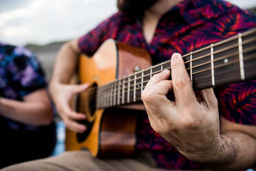 Cropped unerkennbar talentierte machen Musiker in Freizeitkleidung spielen akustische Gitarre und singen Lied, während auf Sandstrand in der Natur im Tageslicht sitzen - ADSF31934