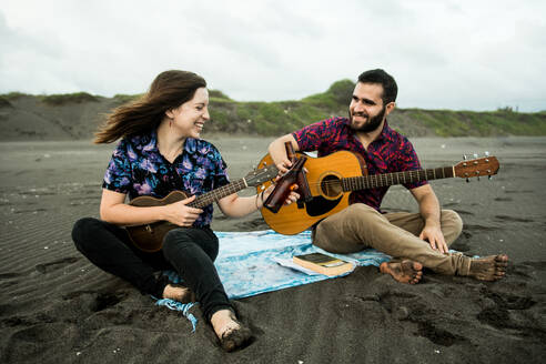 Positives Paar von Musikern, die mit Gitarren sitzen und mit Bierflaschen anstoßen, während sie tagsüber am Sandstrand in der Nähe des Meeres sitzen - ADSF31935