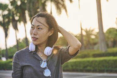 Serious Asian female with modern white headphones looking into distance while standing near road on street of town with green trees - ADSF31937
