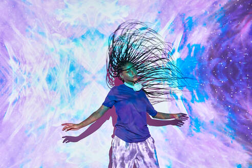 Side view of ethnic female millennial shaking long braided hair while dancing near wall with bright blue and lilac illumination - ADSF31957