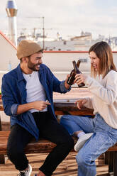 Delighted young friends in casual clothes sitting on wooden bench and clinking bottles of beer while chilling on terrace together - ADSF31959