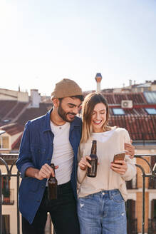 Lächelnder bärtiger Mann mit Bierflasche umarmt positive Freundin scrollt Handy auf Balkon in sonnigen Tag - ADSF31963