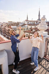 Full body of cheerful friends raising bottles of beer while laughing and dancing on balcony in old city - ADSF31965