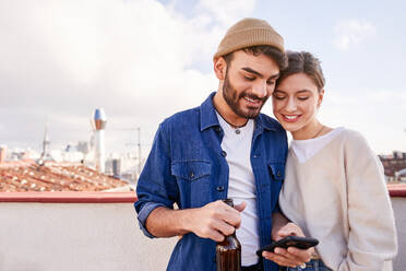 Smiling bearded man with bottle of beer hugging positive girlfriend scrolling mobile phone on balcony in sunny day - ADSF31967