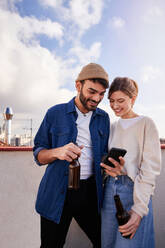 Smiling bearded man with bottle of beer hugging positive girlfriend scrolling mobile phone on balcony in sunny day - ADSF31968