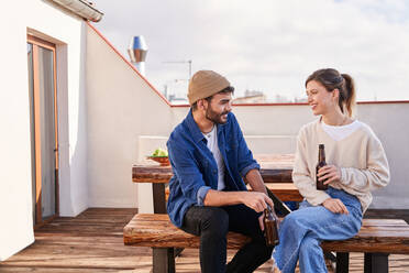 Positive junge männliche und weibliche Mitbewohner, die mit Bierflaschen auf einer Bank sitzen und sich auf der Terrasse angenehm unterhalten - ADSF31969
