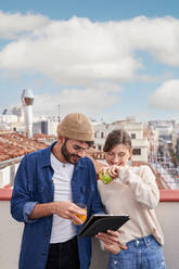 Junger Mann mit einem Glas Orangensaft, der neben einer Mitbewohnerin steht, die auf dem Dach einen gesunden grünen Apfel isst und gemeinsam auf einem Tablet surft - ADSF31974