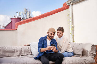 Smiling young woman sitting close to bearded boyfriend and browsing smartphone on couch on terrace - ADSF31983