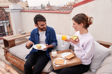 Positive Frau sitzt auf der Couch und isst ein leckeres Sandwich mit Avocado-Scheiben und trinkt ein Glas Orangensaft - ADSF31986