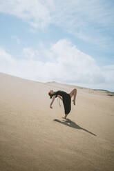 Full body side view of barefoot female dancer outstretching arms while bending back and raising leg in sandy desert - ADSF32015