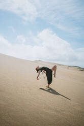 Ganzkörper-Seitenansicht einer barfuß gehenden Tänzerin, die ihre Arme ausstreckt, während sie den Rücken beugt und das Bein in der Sandwüste anhebt - ADSF32015
