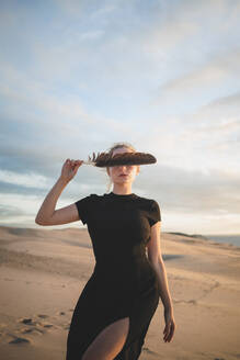 Ernste Frau, die ihre Augen mit einer schwarzen Feder bedeckt und ein Kleid trägt, das auf einer vom Meer umspülten Sanddüne bei Sonnenuntergang steht - ADSF32020