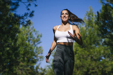 Von unten lächelnd sportliche Frau in Sportkleidung läuft auf dem Lande während Fitness-Workout zwischen grünen Bäumen - ADSF32042