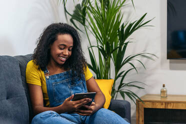 Positive afroamerikanische Frau im Jeans-Outfit sitzt auf der Couch und lächelt, während sie zu Hause auf dem Tablet surft - ADSF32063