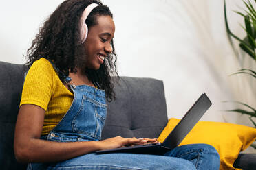 Smiling black woman in denim overalls with headphones sitting on sofa and using laptop at home - ADSF32064