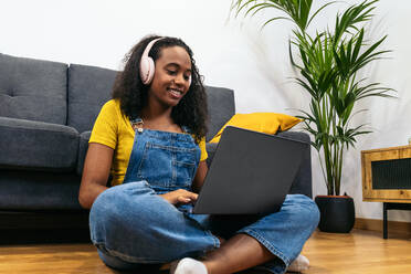 Lächelnde schwarze Frau in Jeans-Overall mit Kopfhörern sitzt auf dem Boden in der Nähe des Sofas und benutzt einen Laptop zu Hause - ADSF32065