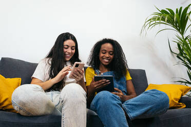 Junge, bunt gemischte Freundinnen in Freizeitkleidung lächelnd auf dem Sofa sitzend, während sie im Wohnzimmer zu Hause auf ihrem Smartphone surfen - ADSF32066