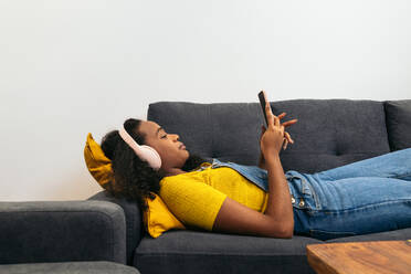 Junge Afroamerikanerin mit lockigem Haar, Kopfhörern und Freizeitkleidung, die auf einem bequemen grauen Sofa liegt und in einem hellen Raum zu Hause auf ihrem Smartphone surft - ADSF32070
