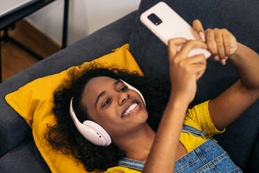 Von oben junge afroamerikanische Frau mit lockigem Haar, Kopfhörern und Freizeitkleidung, die auf einem bequemen grauen Sofa liegt und ein Selfie mit ihrem Smartphone in einem hellen Raum zu Hause macht - ADSF32073