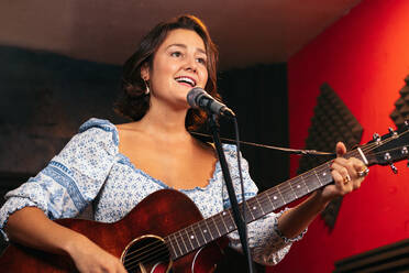 Selbstbewusste Dame mit Gitarre singt in Mikrofon während der Durchführung Song in hellen Club - ADSF32075