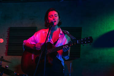 Selbstbewusste Dame mit Gitarre mit geschlossenen Augen singt in Mikrofon während der Durchführung Song in hellen Club mit Neonlicht - ADSF32084