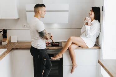 Man looking at woman sitting on kitchen counter - MIMFF00763