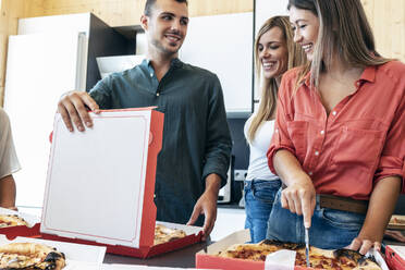 Lächelnde Geschäftsfrau beim Schneiden von Pizza durch Kollegen im Büro - JSRF01721